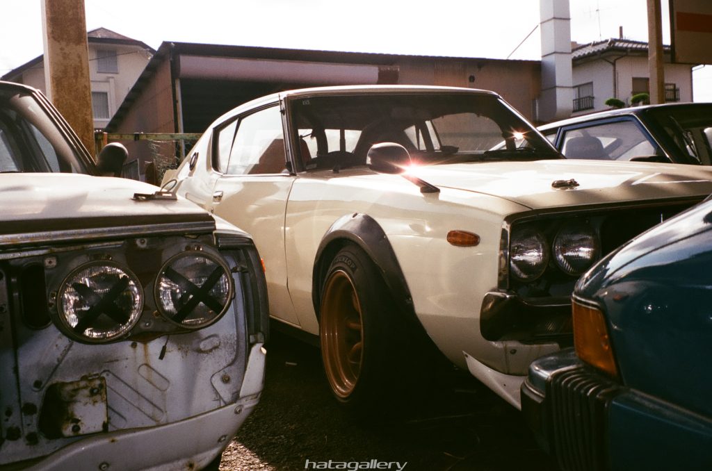 Vintage Japanese sports cars parked outdoors, featuring a white classic model with fender flares and bronze wheels.
