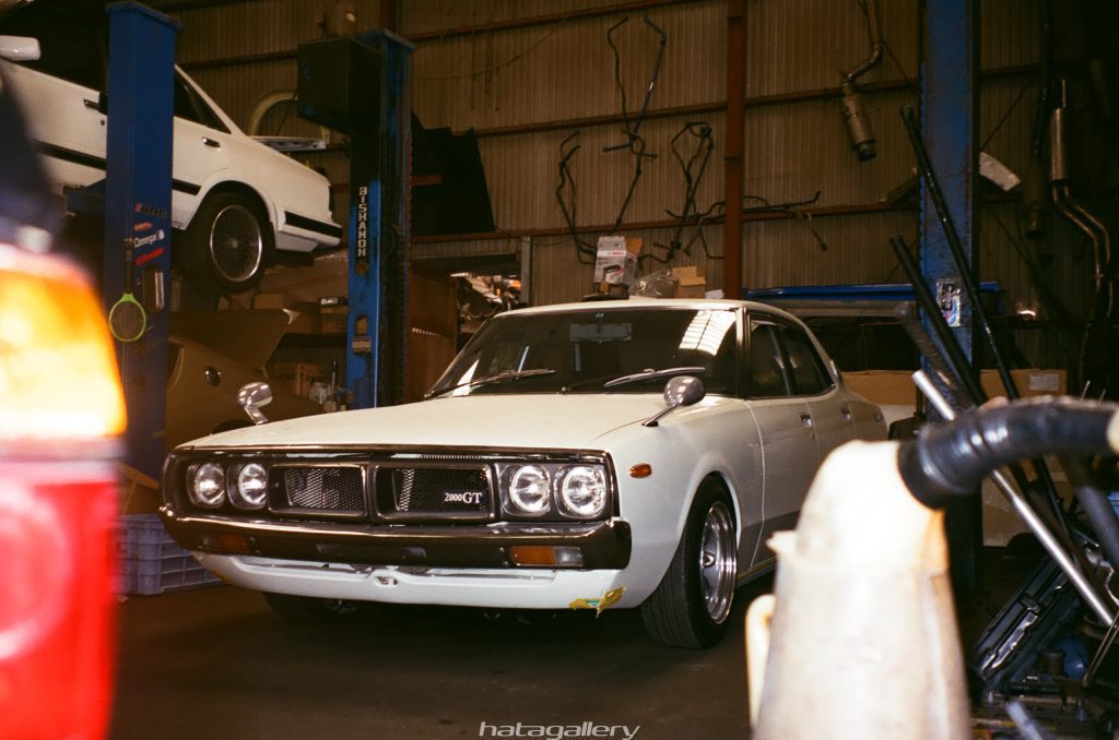 A classic white 2000GT car in a garage with another white car on a lift in the background, hatagallery watermark.