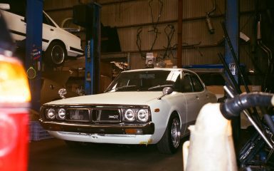 A classic white 2000GT car in a garage with another white car on a lift in the background, hatagallery watermark.