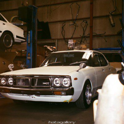 A classic white 2000GT car in a garage with another white car on a lift in the background, hatagallery watermark.