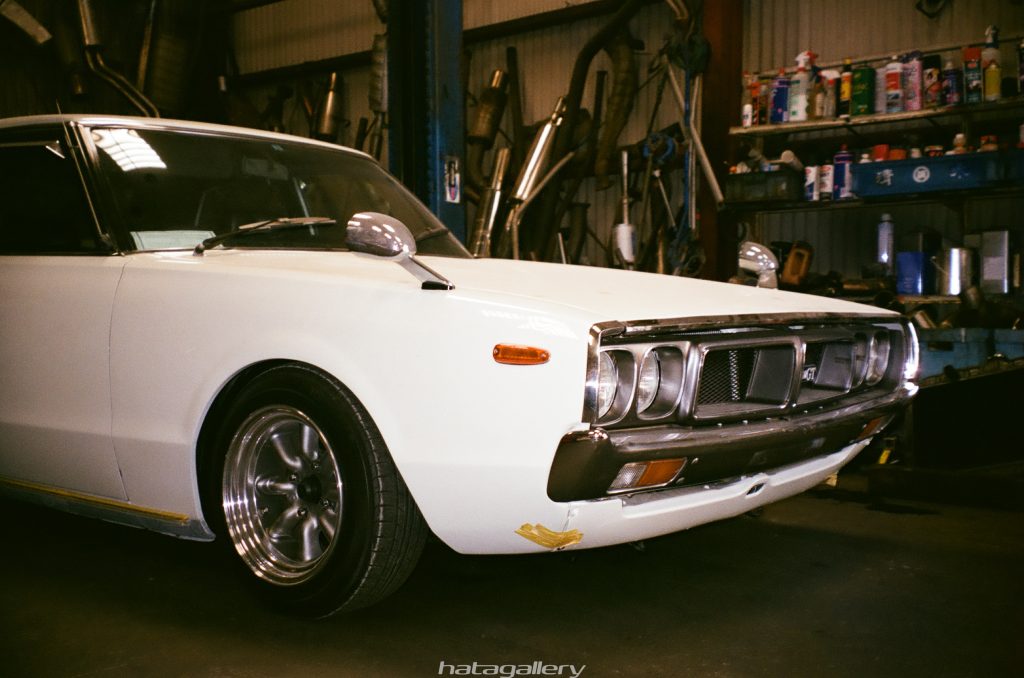 Vintage white car with a GT badge, parked in a garage with various auto parts and tools in the background.