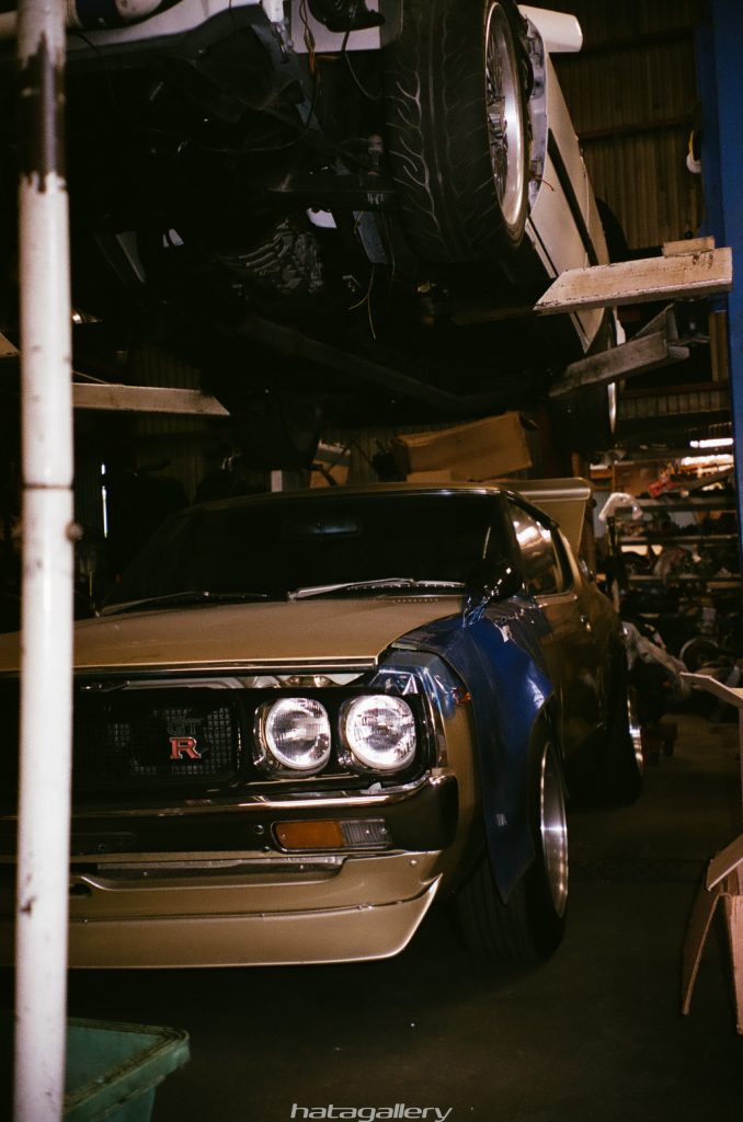 Vintage Nissan GT-R in a garage with another car on a lift, showcasing classic design and automotive restoration.