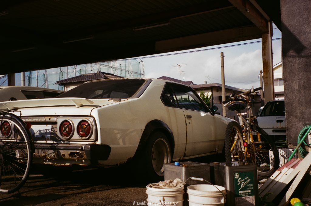 Alt text: Vintage Nissan Skyline car parked under a shed, with bicycles and various items around it, set in a residential area.