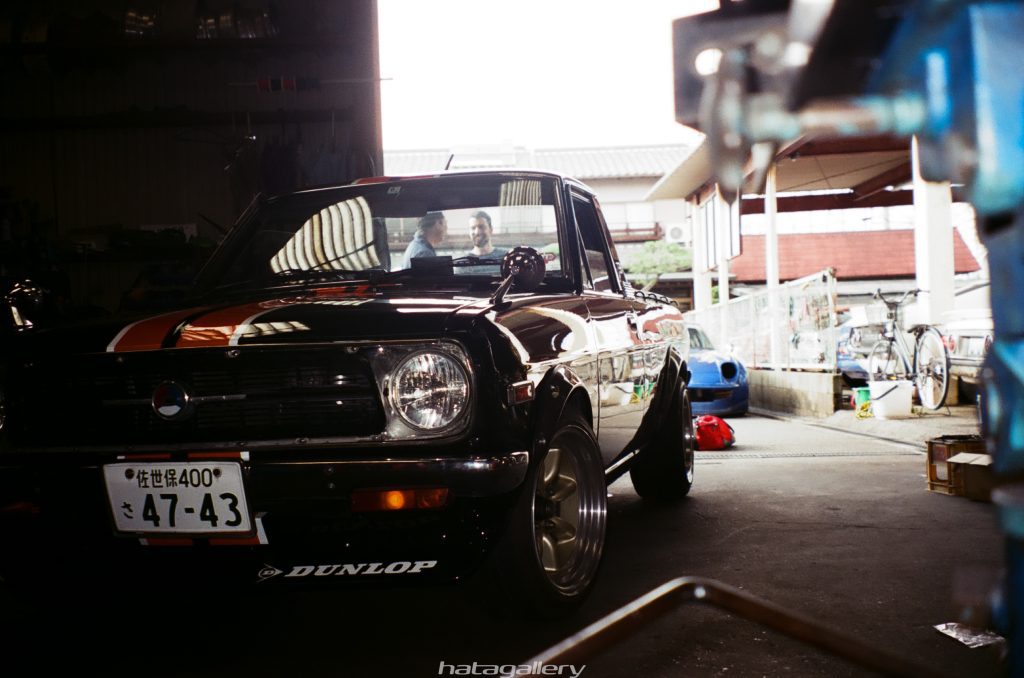 Classic black car in a dim garage with a Dunlop logo and Japanese license plate させ保400 47-43. Two people conversing.