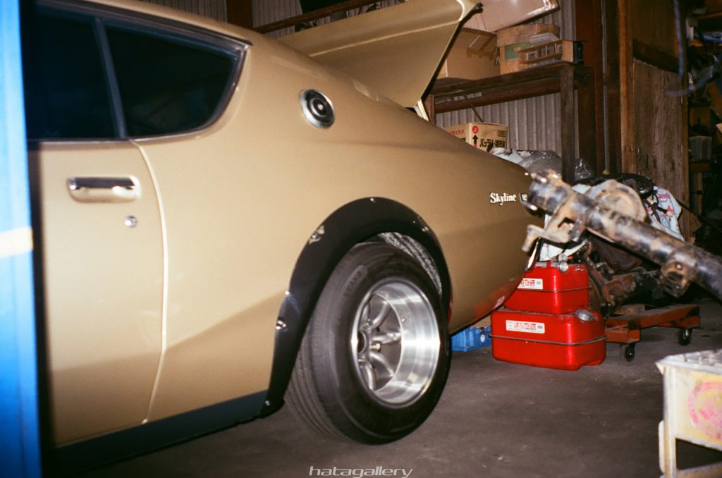Vintage beige Nissan Skyline with trunk open and partially disassembled rear axle in garage filled with tools and car parts.