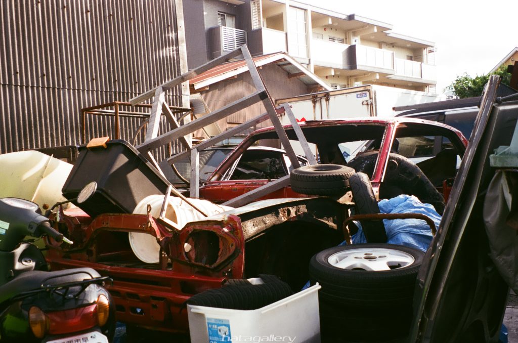 Junkyard scene with red car, tires, and miscellaneous items stacked, translating Japanese sign: 'Nata Gallery.'