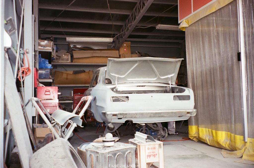 Vintage car undergoing repair and restoration in an auto workshop, with open trunk and surrounding tools and equipment.