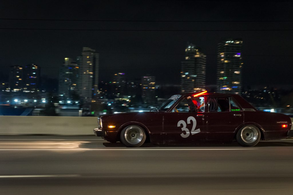 Vintage car with number 32 speeds down a city highway at night, city skyline illuminated in the background.