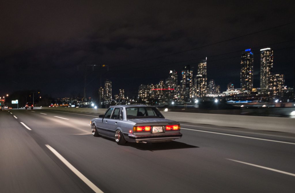Vintage sedan cruises city highway at night, illuminated skyline in background.