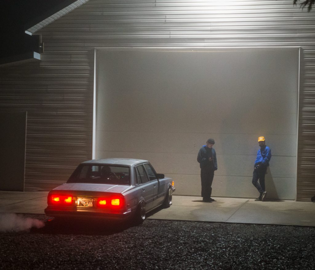 A vintage car with Japanese license plates parked at night near a garage, with two men standing by the garage door.