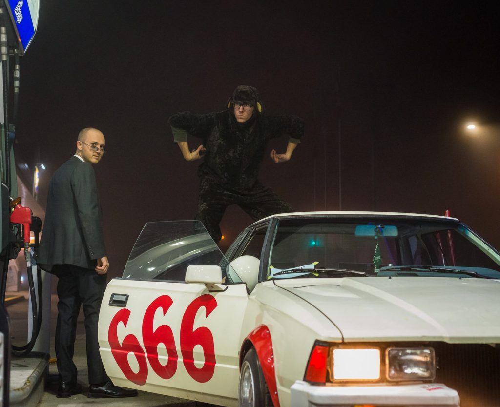 Two men with a car numbered 666 at a gas station at night. One man in a suit, one posing on the car.