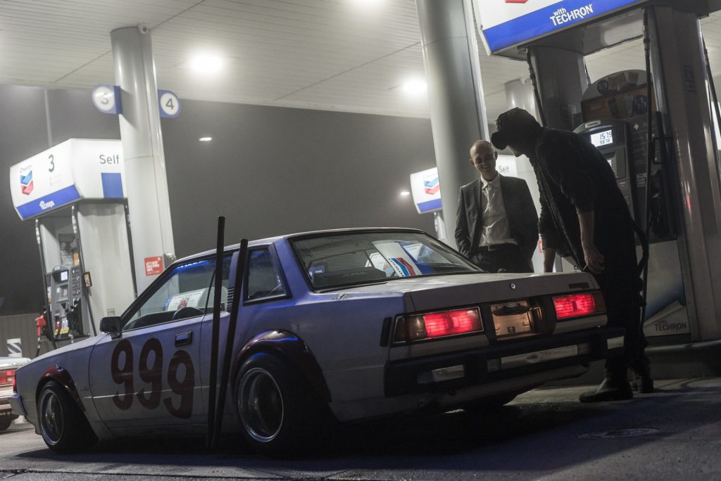 Two people stand by a customized car numbered 999 at a Chevron gas station at night.