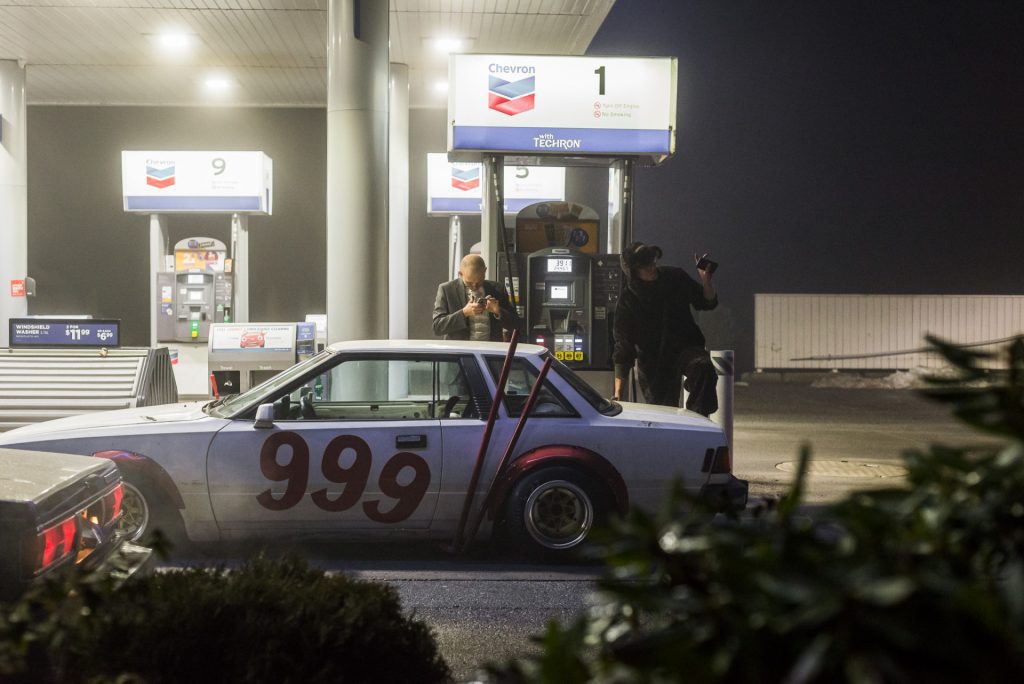 Classic white car with number 999 at Chevron gas station; two men standing near fuel pumps at night, one using phone.