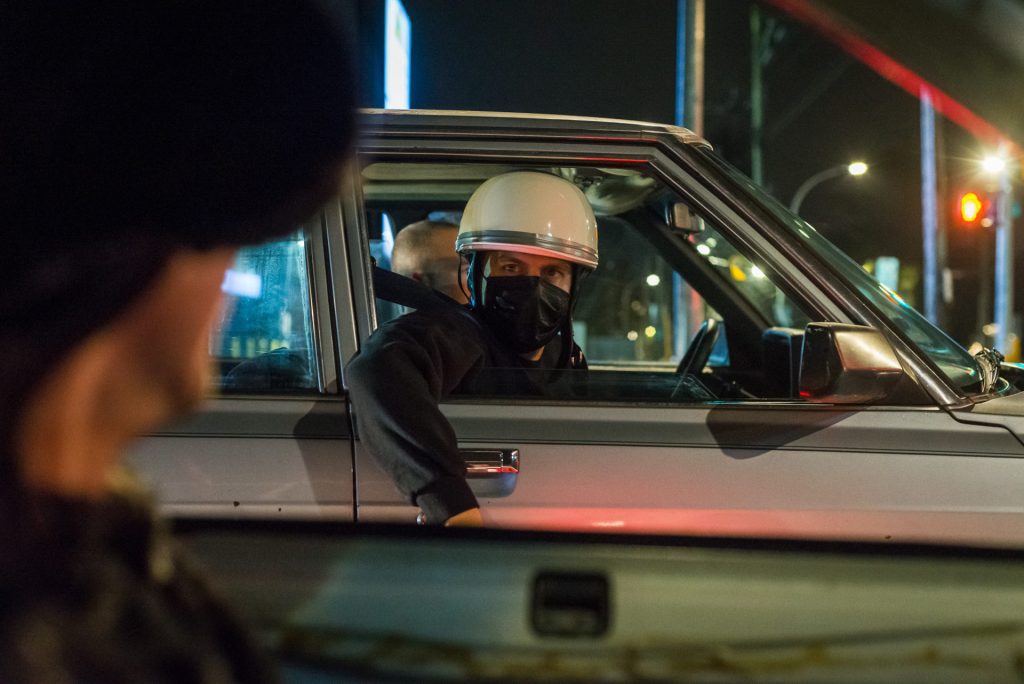 Two masked individuals in cars conversing at night, one wearing a helmet, traffic light in the background.