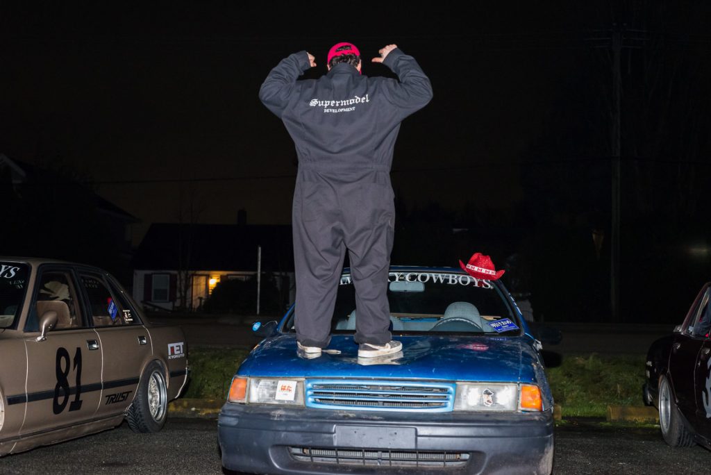 Person standing on a car at night wearing a jumpsuit with Supermodel Development text, cars parked alongside.