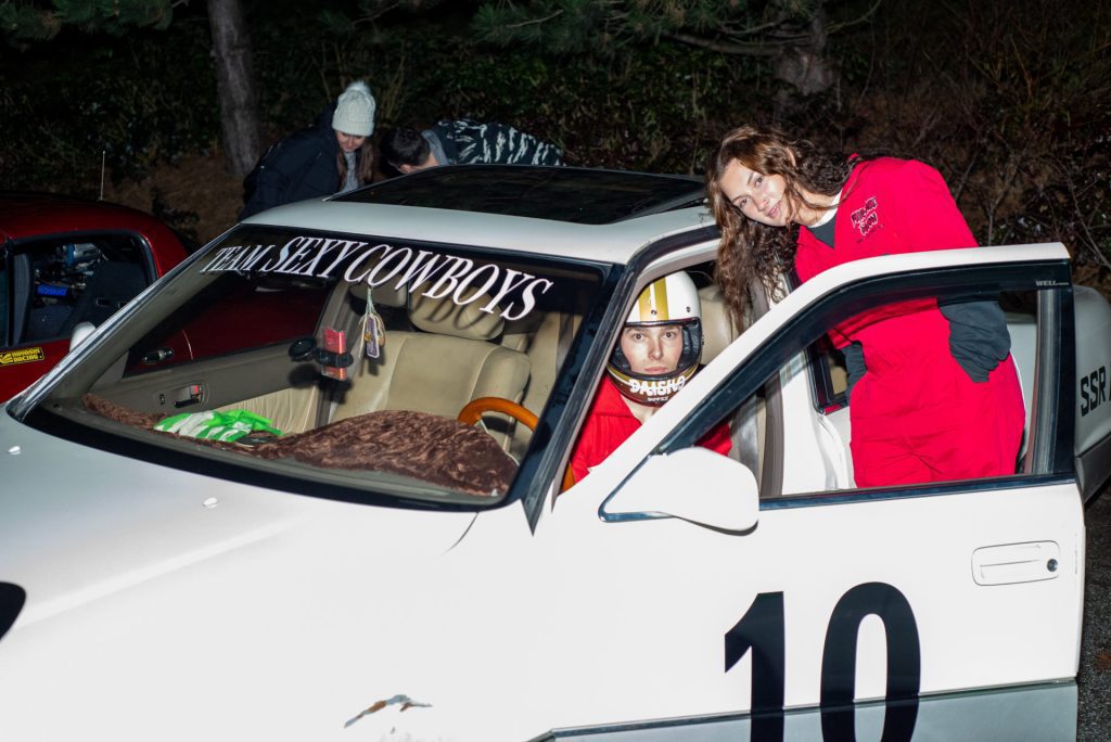 Racecar with Team Sexy Cowboys banner, driver wearing a helmet with Daisho in Japanese, and smiling woman in red suit.