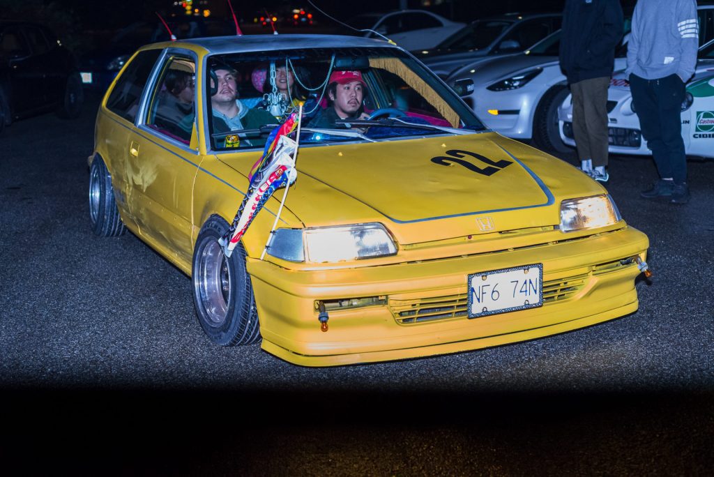 Yellow modified Honda Civic EF hatchback with number 22 and Japanese koinobori flags at night car meet.