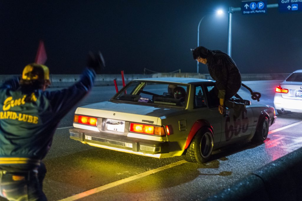Street racing scene at night with a person leaning on a retro car marked '666', another in 'Emil's RAPID-LUBE' jacket.