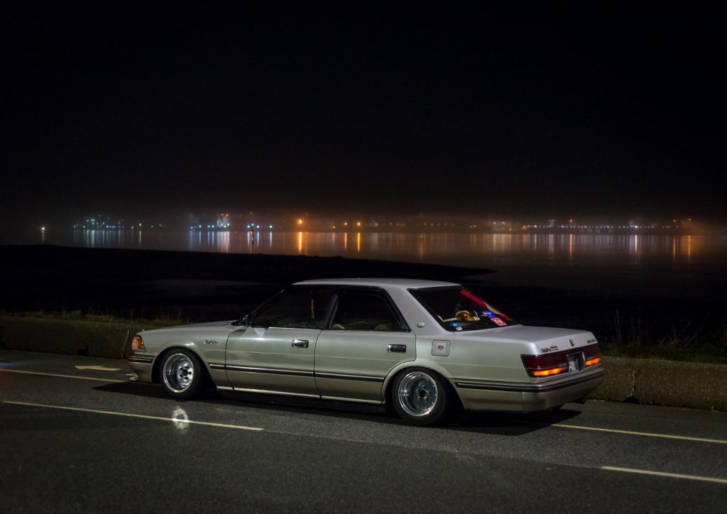 Vintage sedan parked by the waterfront at night, with city lights reflecting on the calm water in the background.
