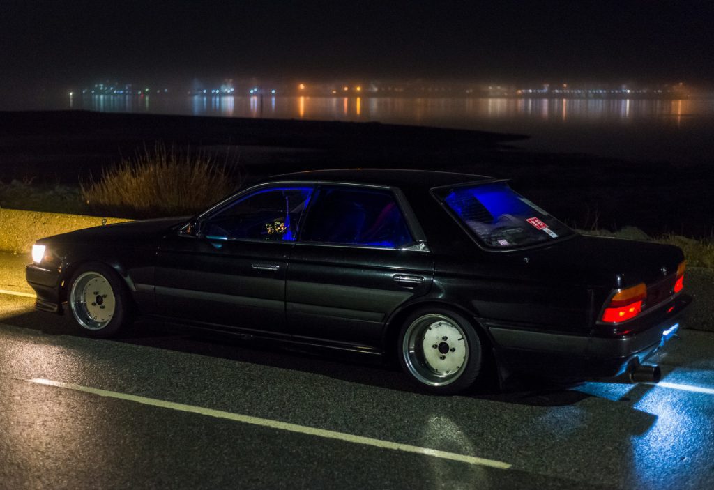 Nighttime view of a modified black Japanese sedan with illuminated interior by the waterfront, reflecting city lights.