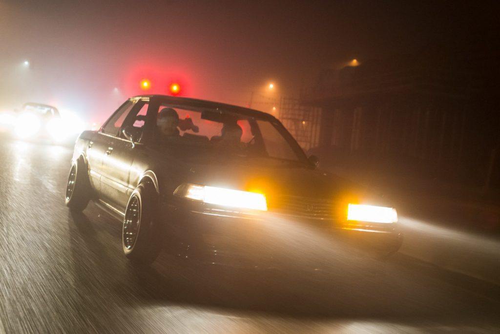 A retro car drives through a foggy night street with bright headlights and blurred streetlights in the background.