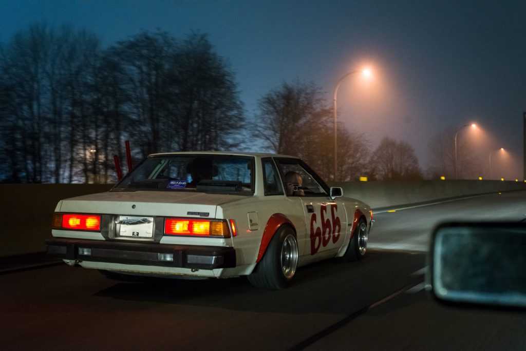 Vintage white car with number 666, driving on foggy highway at night with illuminated taillights and bare trees.