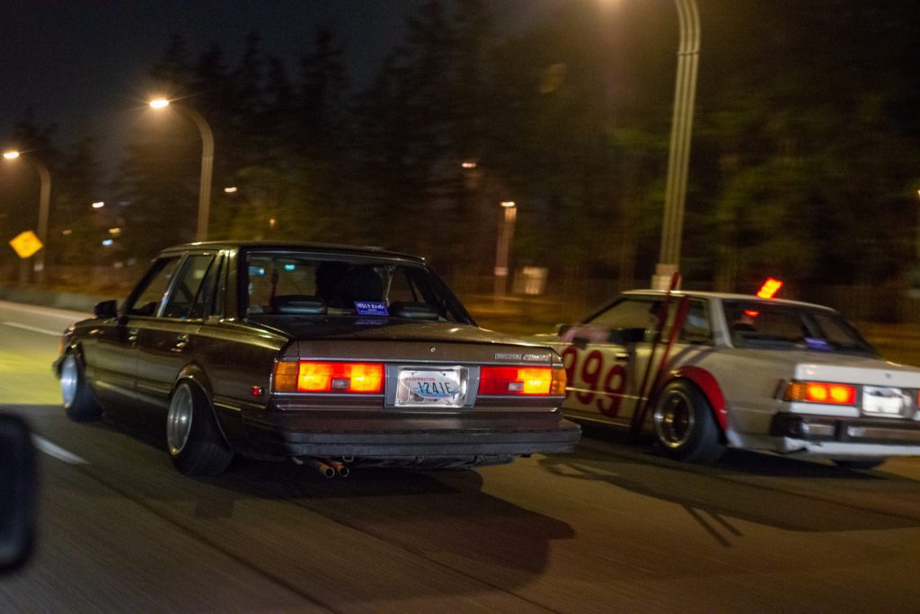 Two vintage cars racing at night on a road in Washington state, one with Japanese number 99 on it.