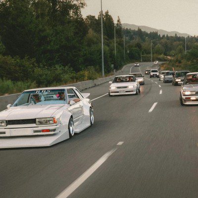 A convoy of modified JDM cars with R.B.S decals on a highway, surrounded by lush greenery, showcasing car culture.
