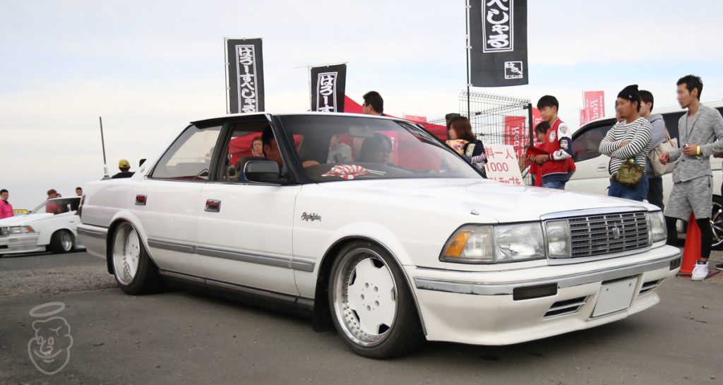 White Toyota Cresta at a Japanese car meet, custom wheels and lowered suspension, surrounded by spectators and banners.