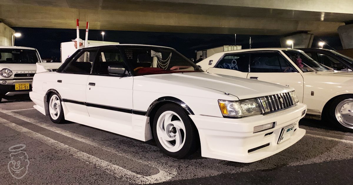 A white classic Japanese car parked under a bridge at night, showcasing its custom body kit and retro design.
