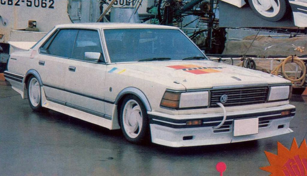 Modified classic white Japanese sedan with racing decals and custom modifications parked near industrial equipment.