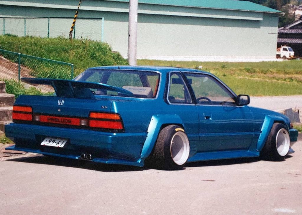 Modified blue Honda Prelude parked with custom wide body kit and spoiler, showcasing Japanese license plate.