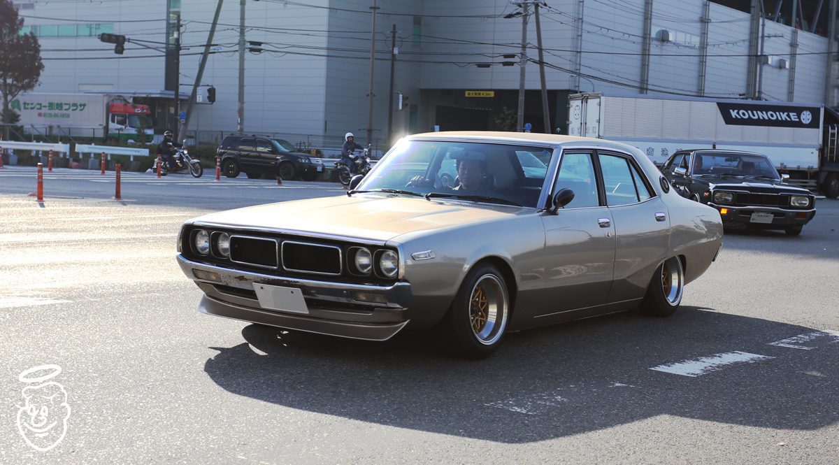 Classic Japanese car driving on a street with several vehicles and the Kounoike truck logo in the background.