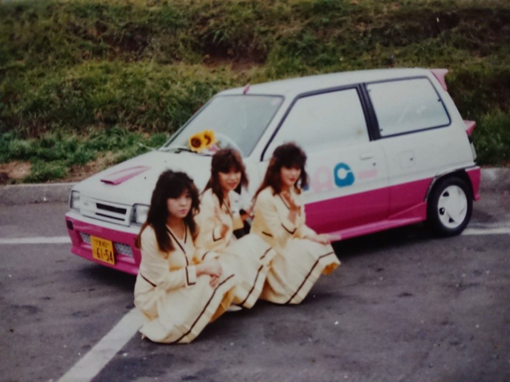 Three women in yellow dresses posing with a pink and white car labeled トラコン (Torakon) in a parking lot.