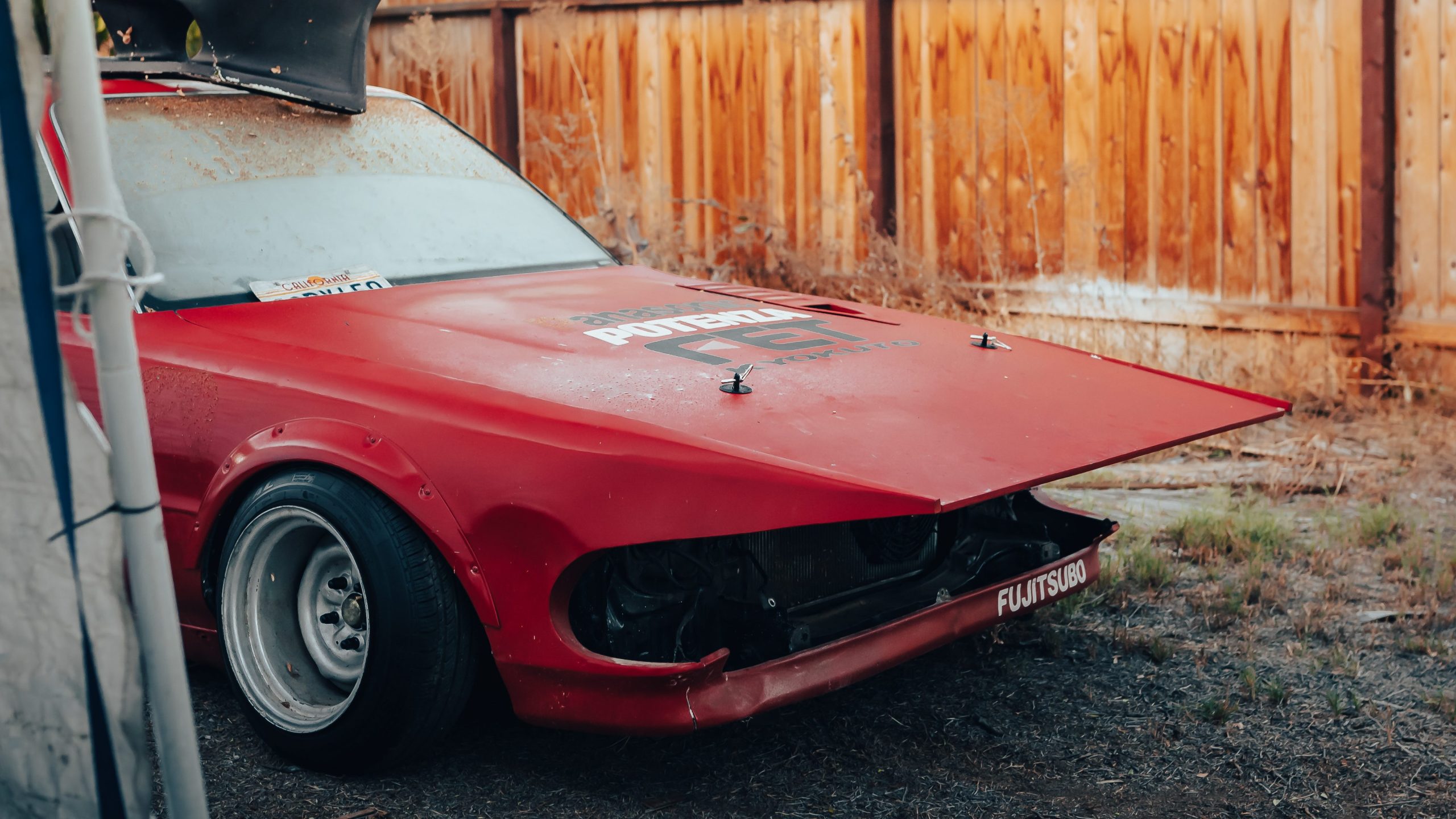 Rusty red classic car with damaged front panel, Fujitsubo decal, and California license plate parked by wooden fence.