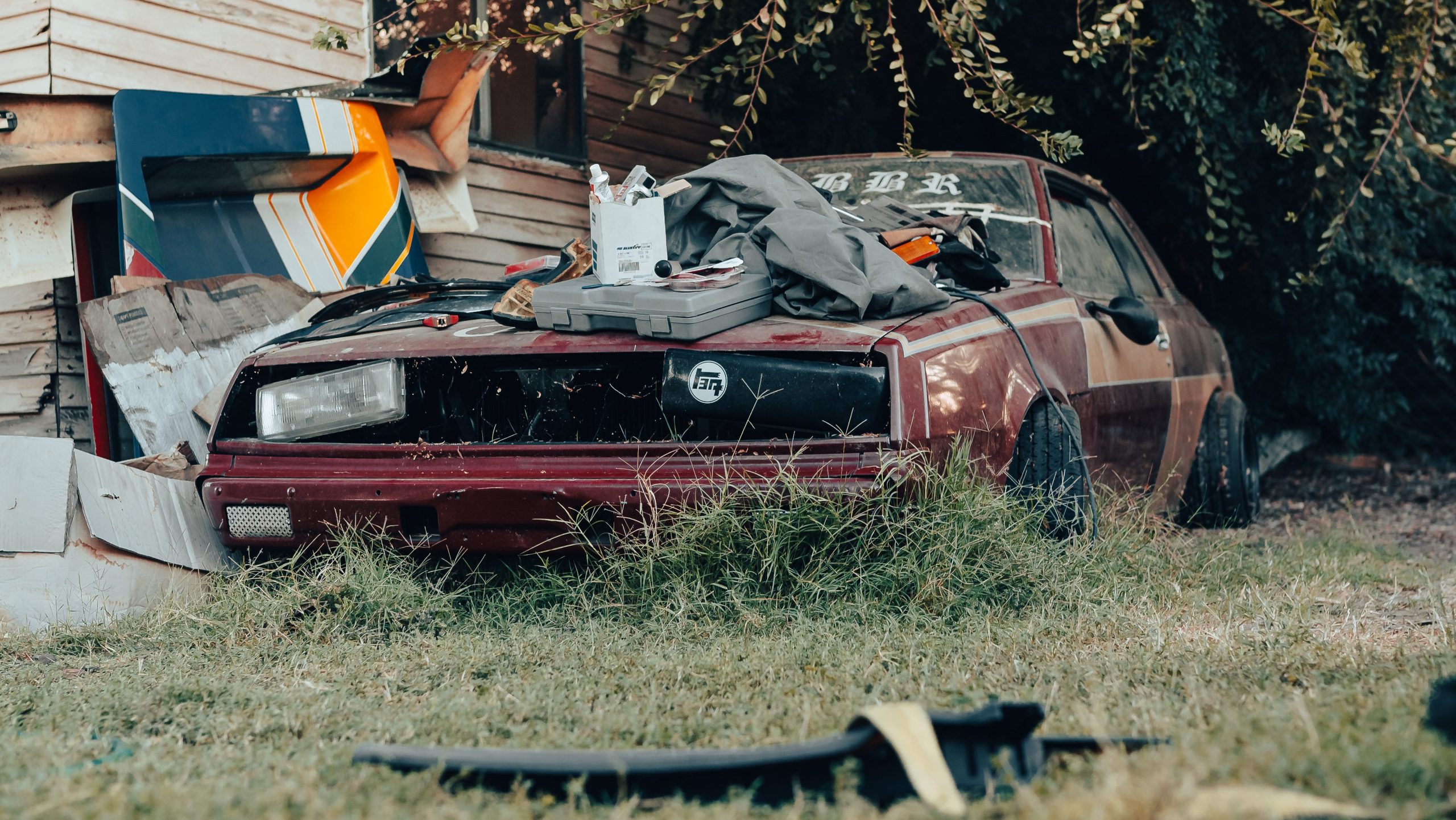 Abandoned red car with tools and clutter on the hood, overgrown grass, and old building in the background.