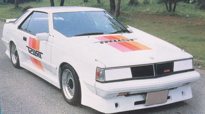 Vintage modified white sports car, Nissan Leopard, with Trust decals and customized wheels in a park setting.