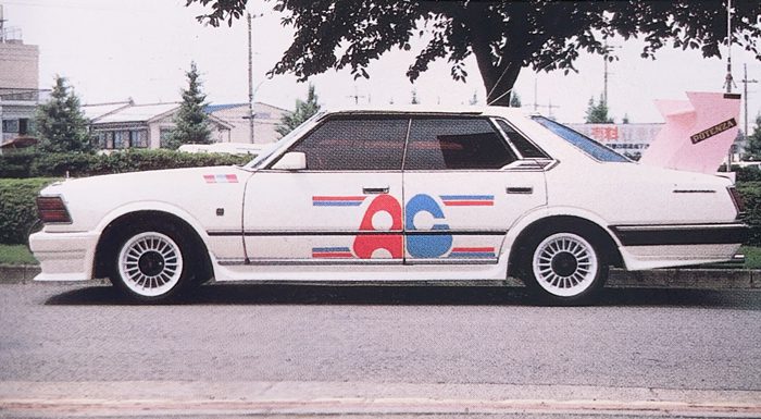 White vintage car with AG racing decals parked on street in Japan, featuring tree and houses in the background.