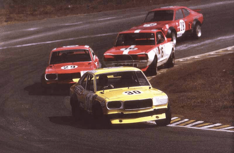 Four vintage race cars competing on a track, led by a yellow car number 30, with cars 29, 51, and 16 following closely behind.