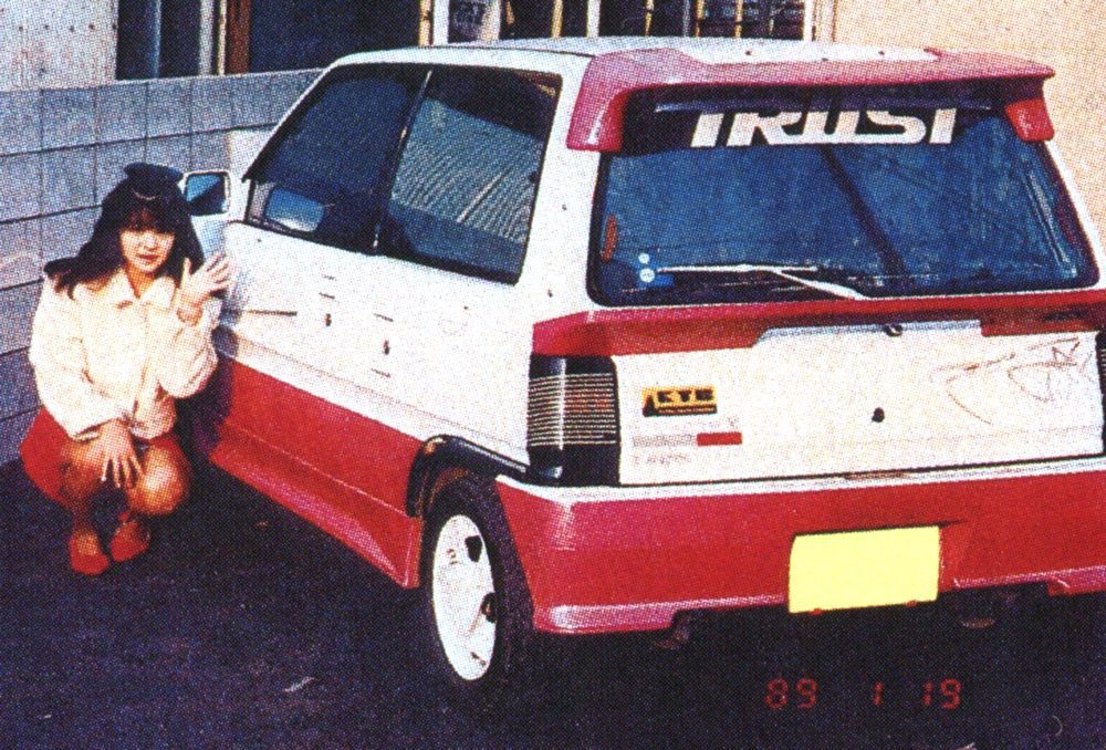 A woman poses next to a vintage Japanese car with a TRUST decal on the rear windshield, dated January 1989.