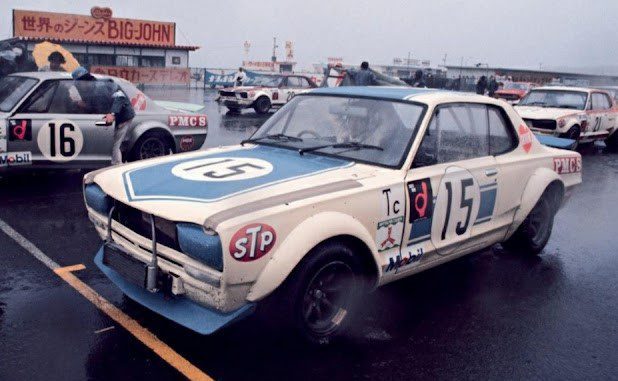 Vintage race cars in a wet pit area with a large Big John sign in Japanese overhead, featuring the number 15 car in foreground.