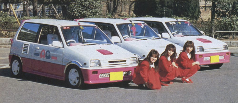 Women in red uniforms pose with three white and pink customized Honda City Turbo II cars showing ACG logo, outdoor setting.