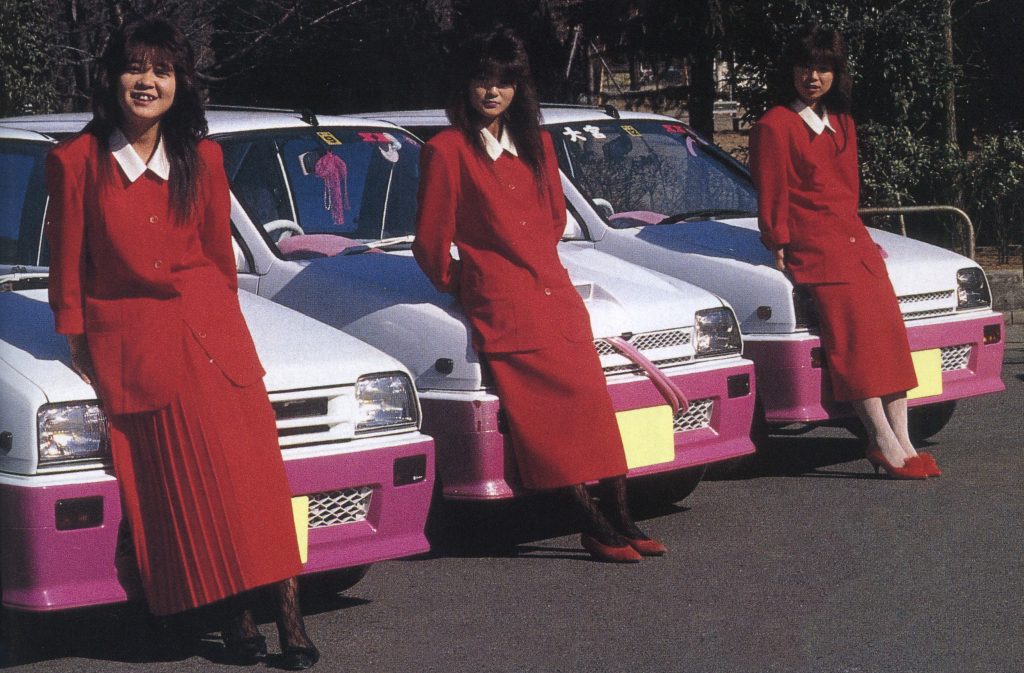 Three women in red outfits lean against white cars with pink accents in Japan. Japanese text translates to no passengers.