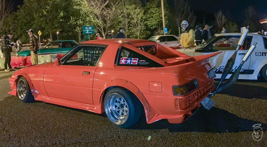 A modified red sports car with oversized exhausts at a night car meet, surrounded by onlookers and other vehicles.
