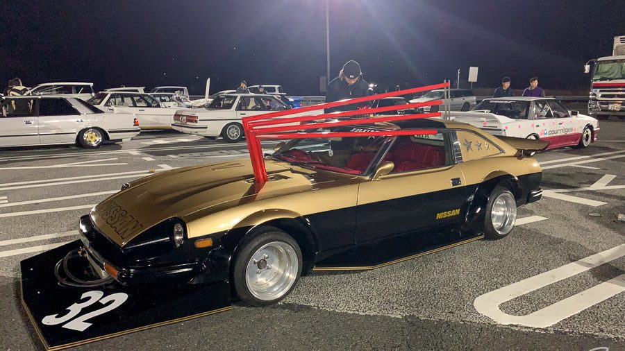 Gold and black modified Nissan car with Japanese characters and number 32 in a nighttime car meet with other classic cars.