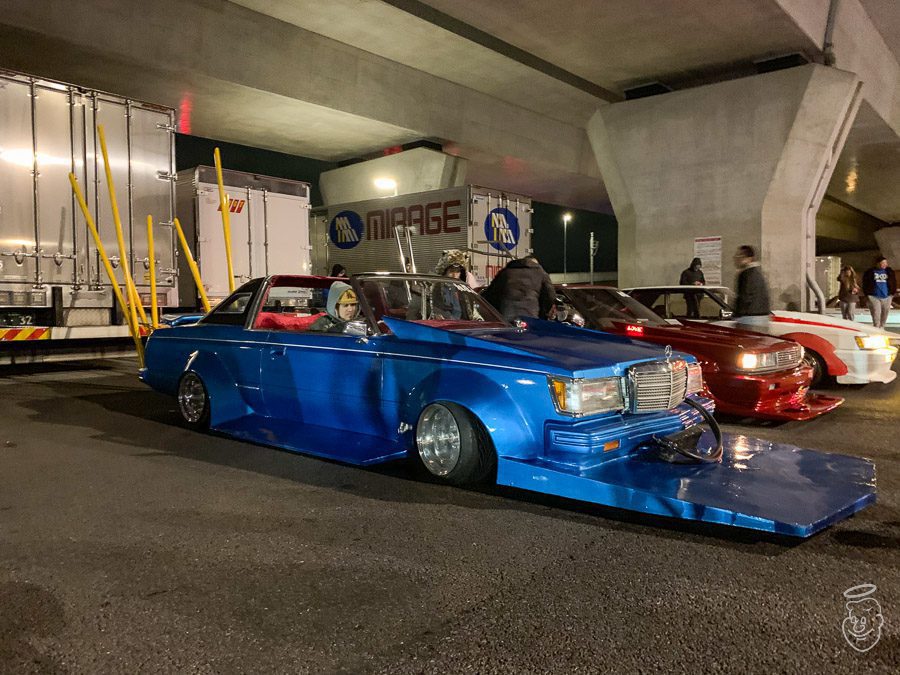A vivid blue lowrider car with a unique body kit, parked under a highway, surrounded by trucks and other modified cars at night.