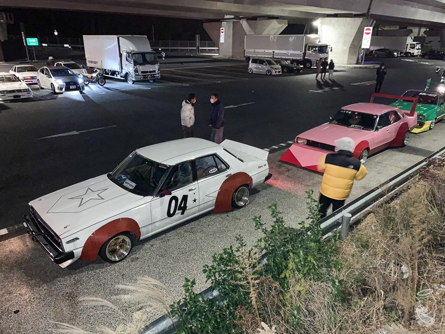 Vintage Japanese modified cars parked under an overpass at night with people standing nearby.