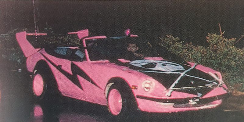 Pink custom sports car with black lightning bolt design and unique hood art, driven at night on a wet road.