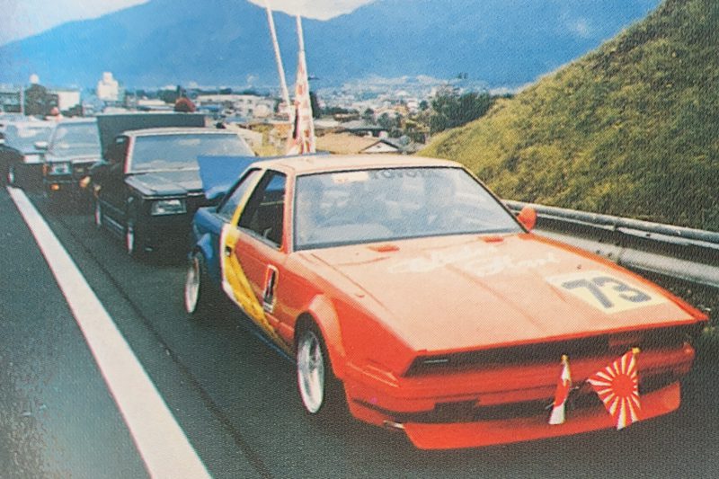Brightly colored sports car with Japanese flags on the highway surrounded by other cars, mountains in the background.