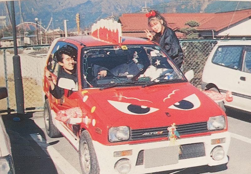Two people with peace signs next to a red car styled with cartoonish eyes on its hood in a parking lot in Japan.